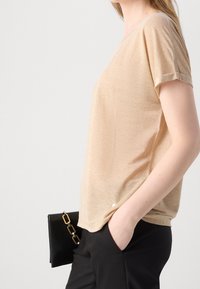 Top de manga corta beige con un corte relajado y un ligero brillo. Combinado con pantalones negros y un bolso negro con un acento de cadena dorada.