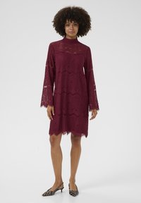 Vestido de encaje burdeos con mangas largas acampanadas, escote alto, dobladillo con forma de onda y patrones intrincados. Combinado con zapatos a rayas en blanco y negro.