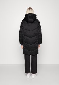 Svart vadderad lång kappa med huva, med chevron-quilting och en slät yta. Modellen bär svarta byxor och vita skor.