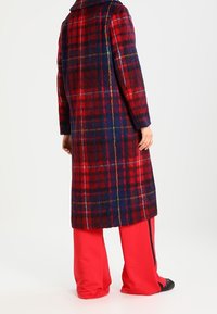 Abrigo de cuadros rojos y azules con un collar ancho, hecho de una tela texturizada y con corte recto, llevado sobre pantalones rojos brillantes.