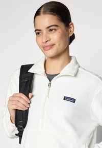 Suéter de forro polar blanco con cremallera de cuarto de vuelta, cuello alzado y logotipo de Patagonia en negro. El modelo sostiene una correa de mochila negra, mostrando la textura suave.
