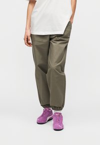 Χαλαρό παντελόνι σε ελιά πράσινο με γυρισμένο τελείωμα στον αστράγαλο, συνδυασμένο με μοβ σουέντ αθλητικά παπούτσια με λευκές λεπτομέρειες και κορδόνια. Απλό λευκό μπλουζάκι.