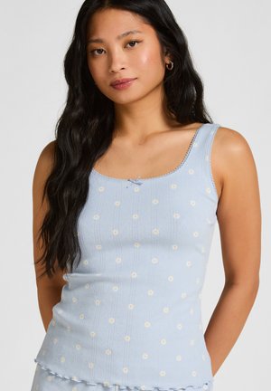 Jonge vrouw met lang zwart haar, gekleed in een lichtblauw hemdje met een patroon van kleine witte madeliefjes, staand tegen een effen achtergrond.