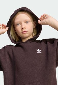 Felpa con cappuccio oversize di colore marrone scuro, realizzata in tessuto morbido, con una grande tasca frontale e il logo Adidas bianco sul petto. Cappuccio indossato.