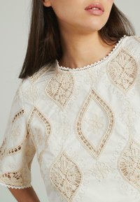 Witte geborduurde blouse met diamant- en bloempatronen, doorschijnende delen, geschulpte halslijn en textuurdetails.