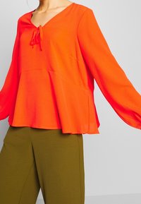 Blusa arancione brillante con scollatura a V, dettagli di lacci al collo e maniche lunghe. Tessuto leggero con finitura testurizzata.