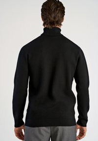 Schwarzer Rollkragenpullover mit einer engmaschigen Textur, geripptem Kragen und langen Ärmeln, kombiniert mit grauen Hosen, von hinten betrachtet.