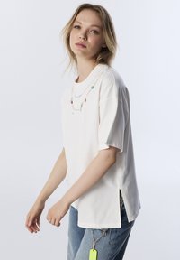 T-shirt bianco in cotone a maniche corte, con ricamo di collana decorativa a catena e spacchi laterali. Abbinato a jeans azzurri.