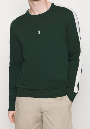 Mand iført mørkegrøn langærmet sweatshirt med hvide striber på ærmerne og beige bukser, stående mod en hvid baggrund.