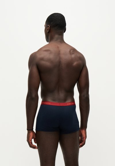 Homme musclé à la peau foncée, debout de dos, portant un boxer bleu marine avec une ceinture rouge Emporio Armani.