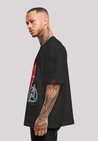 Černé oversized tričko s grafickým potiskem červené postavy a logem rozbitého kruhu. Krátké rukávy a měkká textilie.