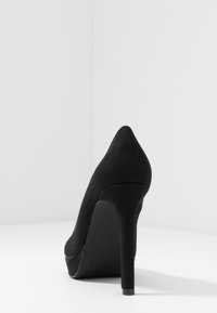 Černá vysoká podpatková botka s špičatou špičkou a hebkou strukturou. Vyznačuje se elegantním designem a stiletto podpatkem, umístěná na bílém pozadí.