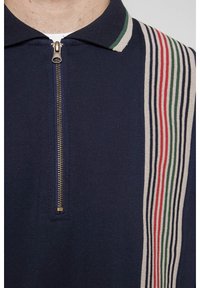 Polo de color marino con cremallera que presenta un patrón a rayas en contraste en rojo, verde y crema a lo largo del hombro. Textura de tela suave.