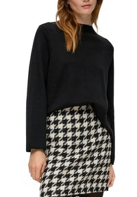 Vrouw in een zwarte lange mouw trui en een zwart-wit houndstooth patroon rok, staand met haar arm ontspannen.