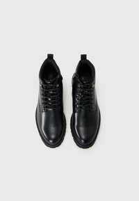 Botines de cuero negro hasta el tobillo con cordones en la parte delantera, punta redonda y suela de goma texturizada. Presenta ojales metálicos y una lengüeta para facilitar su uso.