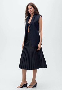 Blazer senza maniche nero abbinato a una gonna midi plissettata nera e sandali infradito neri, che mostrano una silhouette elegante e moderna.
