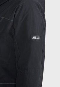 Veste imperméable noire avec une texture lisse, dotée d'un patch logo Aigle proéminent sur la manche et de détails de couture contrastés.