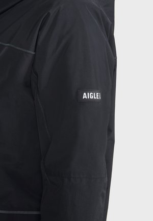 Giacca impermeabile nera con una texture liscia, con un patch del logo Aigle ben visibile sulla manica e dettagli della cucitura in contrasto.