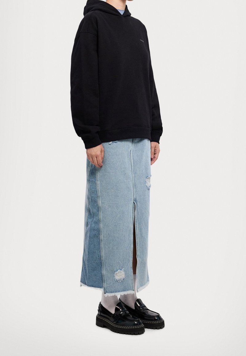 Felpa nera oversize, gonna di jeans blu chiaro con uno spacco frontale e dettagli strappati, abbinata a scarpe nere e calzini bianchi.