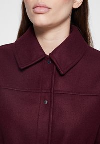 Frau trägt eine dunkelviolette Wolljacke mit breitem Kragen und zwei sichtbaren Metall-Druckknöpfen auf der Vorderseite.