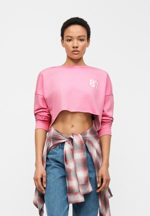 Ung kvinde stående iført en kort pink sweatshirt, blå jeans og en rød og hvid ternet skjorte bundet omkring hendes talje.