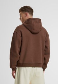 Sweatshirt à capuche marron avec une coupe décontractée, présentant des poignets et un ourlet côtelés. Texture douce, avec une poche kangourou à l'avant.