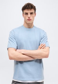 Νεαρός άνδρας με σταυρωμένα τα χέρια, φοράει ένα ανοιχτό μπλε t-shirt BOSS, στέκεται μπροστά σε ένα απλό λευκό φόντο.