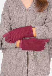 Une femme dans un cardigan gris texturé croise les bras, portant des gants en maille côtelée maroon en fausse fourrure avec des bouts et des poignets en suède.