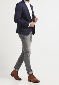 Marineblauwe blazer, wit overhemd, grijze spijkerbroek, bruine leren laarzen. De blazer heeft een smalle pasvorm; de spijkerbroek heeft opgerolde zomen; de laarzen hebben veters en een gestructureerde afwerking.