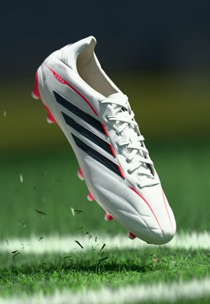 White soccer cleat with black stripes and pink accents, featuring a textured upper, laces, and multiple pointed studs for traction.