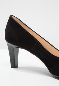 Chaussure à talon haut en daim noir avec un bout arrondi, un intérieur lisse et un talon texturé noir, présentant une silhouette élégante et des lignes épurées.