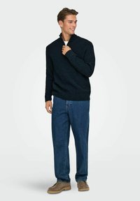 Dunkelblauer Strickpullover mit einem halben Reißverschluss, kombiniert mit lockeren blauen Denim-Jeans und braunen strukturierten Schuhen. Hauchweicher, lässiger Stoffmix.