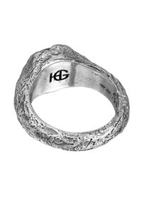 Silver ring med en texturerad yta som liknar naturliga former. Har en ojämn, organisk form och präglad logotyp på insidan.