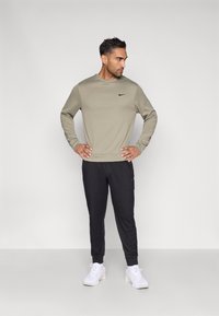 Olijfgroene shirt met lange mouwen en zwart logo, gecombineerd met zwarte taps toelopende broek en witte sneakers. Gladde stof en comfortabele pasvorm.