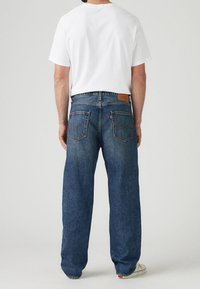 Homme portant un T-shirt blanc, un jean bleu ample et des baskets blanches, debout, tourné de dos sur un fond uni.