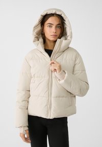 Beige puffjacka med huva, med dragkedjestängning, elastiska ärmslut och quiltad textur, bärs över en svart topp och svarta byxor.