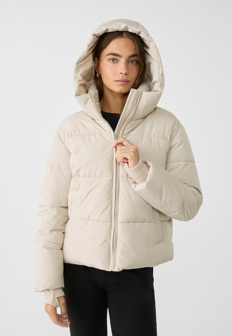 Beige puffjacka med huva, med dragkedjestängning, elastiska ärmslut och quiltad textur, bärs över en svart topp och svarta byxor.