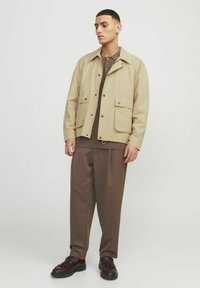 Beige yderjakke med spidse krave, to store frontlommer og trykknaplukning, båret over en brun skjorte med krave og brune bukser.
