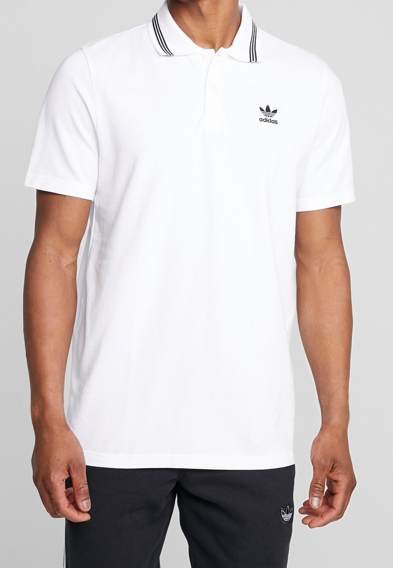Man som bär en vit Adidas pikétröja med svarta ränder på kragen och svart Adidas-logotyp på bröstet, ihop med svarta byxor med logotyp.