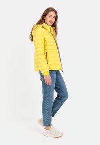 Żółta kurtka puchowa z kapturem, pionowymi przeszyciami, zamek błyskawiczny, boczne kieszenie i beżowe akcenty. Model w niebieskich dżinsach i sneakersach.