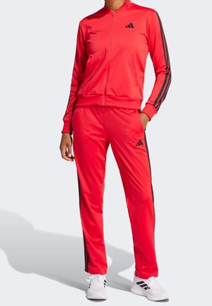 Personne portant un survêtement Adidas rouge avec des bandes noires et des baskets blanches, debout avec une main dans la poche devant un fond uni.