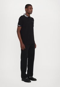 Black polo shirt with striped collar and black accents. Straight-cut black trousers. Model stands facing left, wearing black shoes.