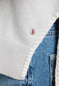 Grauer Strickpullover mit gerippter Textur, ausgestattet mit einem weißen Saum und einem sichtbaren silbernen Knopf mit rotem Emblem. Kombiniert mit blauen Jeans.