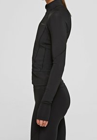 Chaqueta deportiva negra hecha de un material elástico y suave. Presenta un cuello alto, diseño ajustado y agujeros para los pulgares en las mangas largas.