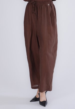 Løse brune teksturerede bukser med elastisk talje og snøre, båret med sorte spidse høje hæle på en lys baggrund.