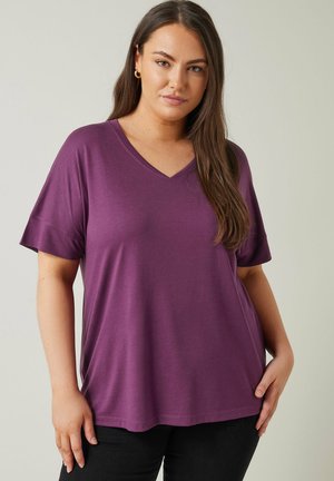 Fioletowa koszulka typu V-neck z krótkim rękawem, wykonana z miękkiej tkaniny. Charakteryzuje się luźnym krojem i gładką teksturą, zestawiona z czarnymi dżinsami.