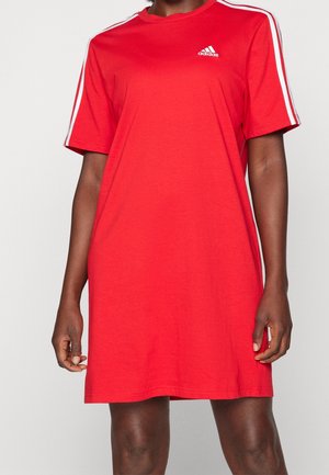 Personne portant une robe T-shirt rouge Adidas avec des bandes blanches sur les manches et un logo sur la poitrine, debout devant un fond uni.