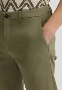 Olivgrüne Hose aus glattem Stoff; mit Knopfverschluss, Seitentaschen und taillierter Passform mit klaren Linien.