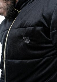 Zwarte corduroy jas met een gequilt ontwerp, ritssluiting en een geborduurd rozenembleem op de linkerborst. Zacht van textuur.