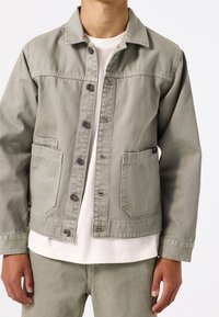 Giacca di jeans verde chiaro con chiusura a bottoni, due tasche frontali, colletto classico e orlo leggermente accorciato. La texture è liscia e resistente.